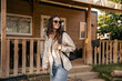 Modern young caucasian brunette woman spends leisure time outdoors near country house. Girl wears sunglasses, shirt, jeans and backpack. Mood, lifestyle, concept.