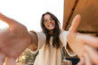 Joyful young caucasian woman in good mood reaches for camera relaxing outdoors. Brunette smiles, wears glasses and sweatshirt. Positive emotions concept