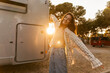 Happy young caucasian woman looking at camera spends vacations in camp at sunset. Brunette waving her arms, wears shirt and jeans. Mood, lifestyle, concept.