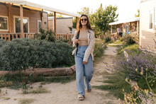 In Full Growth, Nice Young Caucasian Girl Walks Among Cottages On Vacation In Spring. Brunette Wears Sunglasses, Tank Top, Jeans, Slippers And Backpack. People Sincere Emotions Lifestyle Concept.