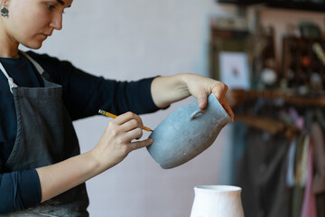 Poster - Young serious skilled woman artisan with clay blue vase and pencil painting handmade earthenware while standing by workplace. Creative craft concept. Focused craftswoman makes sketches on ceramic jug.