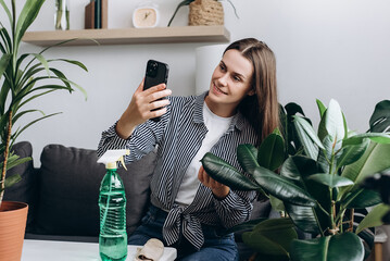 Smiling cute young woman florist blogger being photographed with plants in pot on mobile phone camera. Home plant breeding, gardening, housewife, working online social media influencer concept