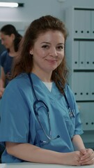 Wall Mural - Vertical video: Portrait of woman nurse sitting at desk in doctors office, looking at camera. Medical assistant with health care expertise working on consultation appointment on computer. Clinical