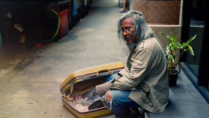 Wall Mural - A homeless old Asian man with his big yellow bag is sitting on the ground in a narrow alley and opening his bag to find something to eat. Homeless people who do not have home to sleep on streets