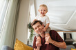 © Arsenii - Laughing father giving his kid piggyback ride on shoulders, baby boy pulling daddy's hair, father and son spending time together on sunday morning in living-room. Dad playing with his toddler boy
