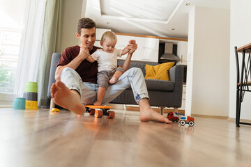 Wall Mural - Young father playing with his little baby boy in living-room, holding his hands while kid trying to stand on skateboard. Active games at home. Carefree childhood, happy fatherhood. Male babysitter