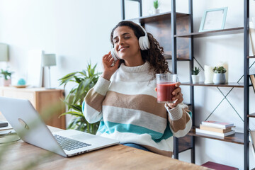 Wall Mural - Pretty young woman listening music with headphone while drinking detox juice working at home.