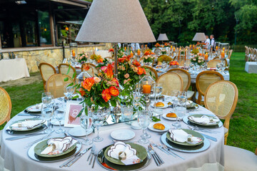 Beautiful decoration of a wedding banquet with a meal in the green garden in white colors. Very stylish setting with tropical flowers in a vase, glass candlestick set, napkins, plates, glasses cutlery