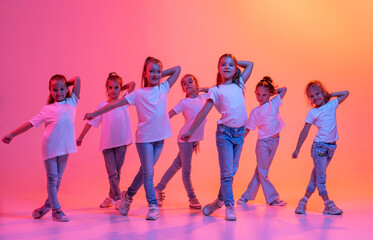 Wall Mural - Group of children, school age girls in sportive casual style clothes dancing in choreography class isolated on pink background in yellow neon light.