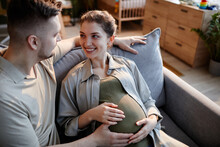 Happy Pregnant Woman Sitting On Sofa With Her Husband And Smiling While He Stroking Her Belly
