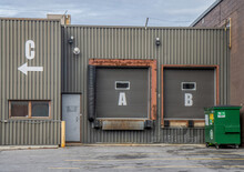 Unoccupied Loading Docks A, B, C On A Low Industrial Building With Green Vertical Metal Siding, Barbed Wire On Roof, Daytime, Nobody