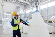 Young Male Engineer Looking Upwards At Huge Hook Where White Sack With Raw Material Hanging While Controling Lifting Process