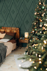 Bedroom decorated for Christmas. Bedroom interior with green Christmas tree decorated with emerald and bronze balls. View through blurred bokeh from garland lights. Christmas interior. Nobody