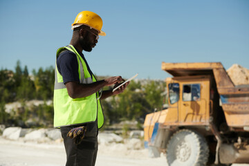 Poster - Side view of young black man in workwear and safety helmet using tablet while looking through online manual guide against truck