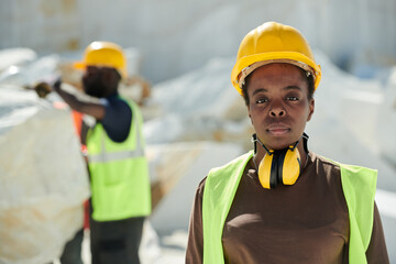 Young serious African American female worker of marble quarry in uniform and protective helmet looking at camera outdoors