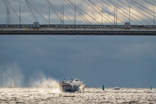 A High-speed Passenger Hydrofoil Boat Passes Under A Cable-stayed Bridge In Sunny Weather, Dark Storm Clouds In The Background