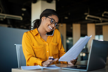 corporate communication. happy black female manager working with papers and talking on mobile phone