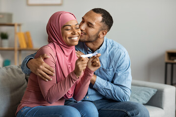 Happy black islamic spouses holding positive pregnancy test and kissing at home