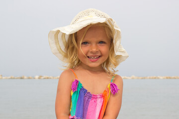 Little blond girl smiling with the sea on background,positive portrait of european toddler,adorable kid on the beach in colorful rainbow dress and hat,cute laughing child looking forward.