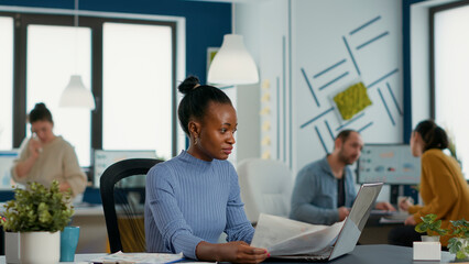 Poster - african american startup employee holding business papers with charts looking at laptop screen compa