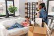 © Syda Productions - moving, people and real estate concept - happy smiling woman unpacking box and her friend resting on sofa at new home