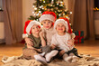 © Syda Productions - christmas, winter holidays and childhood concept - happy little girl and boys in santa hats hugging on floor at home