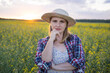 © olga_sova - A beautiful middle-aged farmer woman in a straw hat and a plaid shirt stands in a field of flowering rapeseed in the sunlight