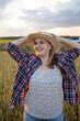 © olga_sova - A beautiful middle-aged farmer woman in a straw hat and a plaid shirt stands in a field of golden ripening wheat during the daytime in the sunlight