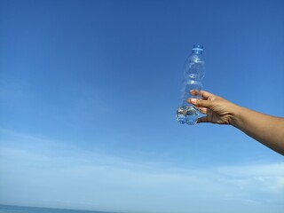  Bottle of mineral water. Person showing transparent bottle with half of water in hand against blue sky background. Copy space. Healthy lifestyle concept. Recycle plastic bottle.