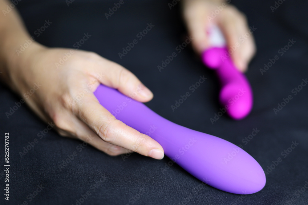 Sex toys in female hands on black bed sheet. Woman on a bed with red  silicone dildo and purple vibrator Stock Photo