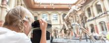 Cute Little Adorable Curious Blond Kid Girl Watching AR Reality Binoculars Exploring Dinosaur Skeleton Exposition At German Nature History Museum. Children Weekend Holiday Place To Visit Concept