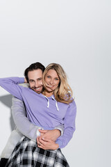 Wall Mural - happy man hugging cheerful woman in purple sweatshirt while looking at camera on grey