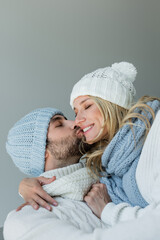 Wall Mural - bearded man kissing cheek of happy blonde woman in knitted hat isolated on grey