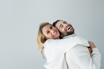 Wall Mural - positive blonde woman in white knitted sweater hugging bearded man isolated on grey