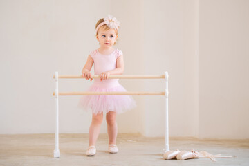 A cute blue-eyed baby ballerina in a pink leotard and tutu standing near a ballet barre. Dream concept of becoming a ballerina.