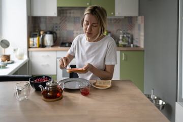 Middle-aged unkempt woman spreads jam on toast having breakfast alone. Mature blonde female eats healthy food in morning thinking about own lifestyle. Lady prepares humble breakfast in late morning