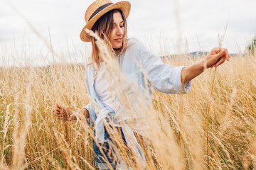 Wall Mural - Portrait of young woman walking among high grasses in summer meadow wearing straw hat and linen shirt.