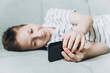 © Lyubov - Teenage boy lying on couch using smartphone and playing video games on internet online at cozy home. Child holding mobile phone and looking at screen. Social media
