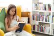 © Alisa Chikov - A young girl is reading a book while sitting in a bright yellow armchair in a public library. The concept of home education, recreation, mindfulness. The girl is wearing light clothes , side view .
