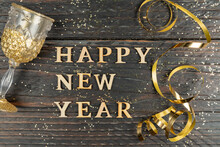 Happy New Year Greeting Card. Glass Of Champagne And Serpantine Of Gold Color On Wooden Table With Scattered Confetti.