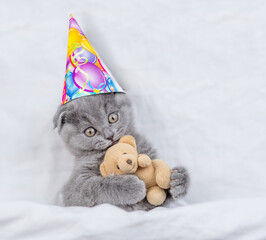 Poster - Cute kitten wearing birthday cap hugs toy  bear under blanket on a bed at home. Top down view
