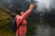 © Yoel Garay - Chica con una chaqueta rosa con una mochila parada junto al lago en las montañas y tomando selfies