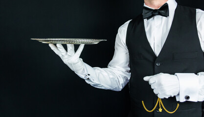 Wall Mural - Portrait of Waiter or Butler in Black Vest and White Gloves Holding Silver Serving Tray on Black Background. Concept of Elegant Service and Hospitality.