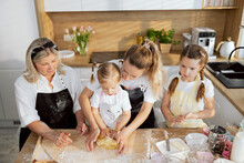 Older Daughter Wiping Hands From Flour While Preschooler Daughter Kneading Dough With Young Mother. Elderly Grandmother Watching Process Comanding.
