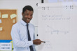 © Seventyfour - Waist up portrait of young black teacher explaining math problem to class and pointing at flipchart
