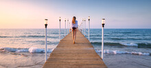 Happy Young Woman In Bikini Walking On Wooden Pier On The Sea At Sunset In Summer. Lifestyle And Travel. Beautiful Girl On Jetty, Sandy Beach, Blue Water With Waves, Sky In The Evening. Tropical 
