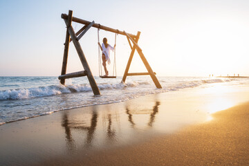 Wall Mural - Happy young woman on wooden swing in water, beautiful blue sea with waves, sandy beach, reflection in water, golden sky at sunset. Summer in Side, Turkey. Girl ride on a swing on sea coast. Travel