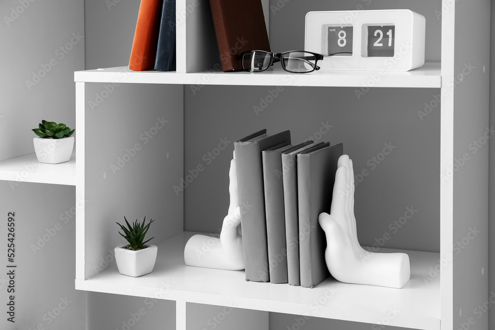 Bookcase with holder, clock and flowerpots near light wall