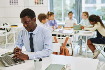 Wall Mural - Portrait of young black teacher using laptop in classroom with group of children in background