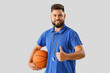 Handsome PE teacher with ball showing thumb-up on light background